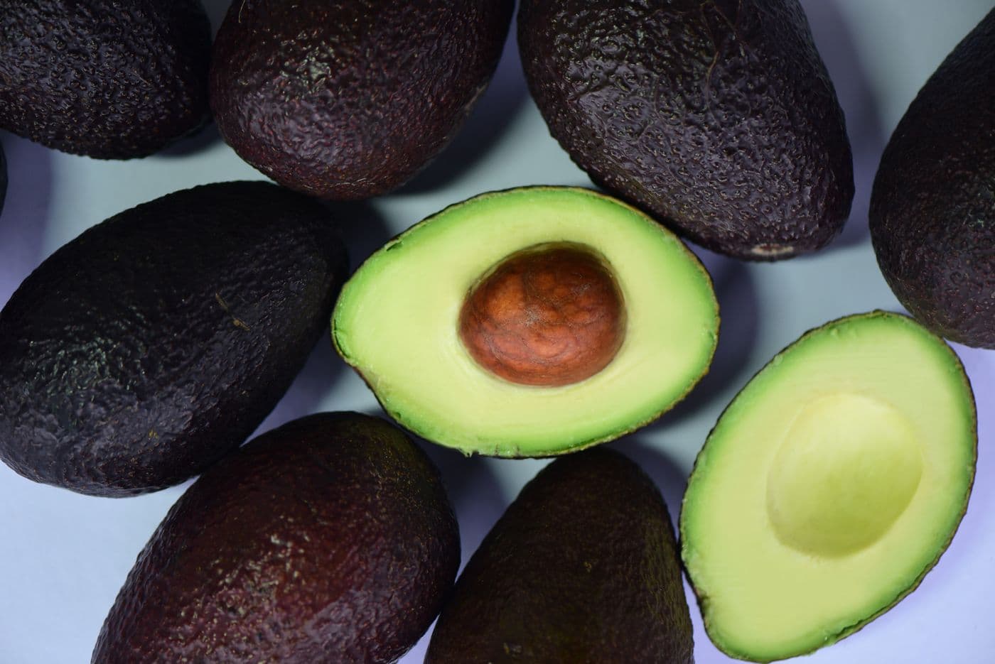 Several dark-skinned avocados with one cut in half showing green flesh and a brown stone, shot from above