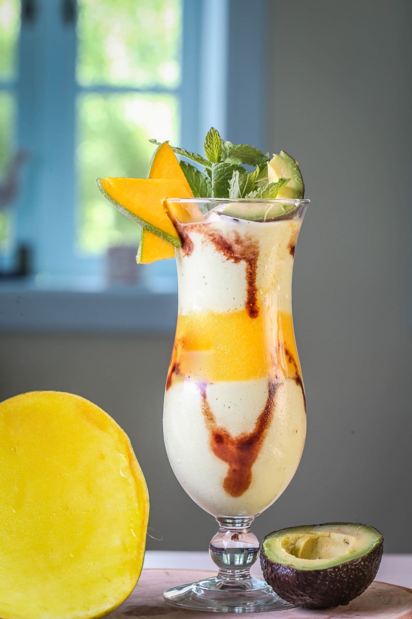 A tall layered mango-avocado smoothie garnished with mint, with a mango half and an avocado half beside it on a wooden table