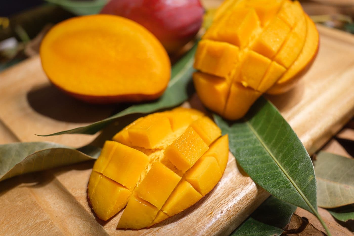 A ripe mango halved and scored into cubes showing orange-yellow flesh on a wooden board with green mango leaves